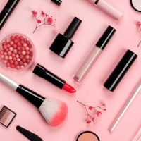 Various cosmetic accessories for makeup and manicure on trendy pastel pink background with red flowers. Blush, brush, eye shadow, mascara, perfume, lipstick, nail Polish. Skin care products.